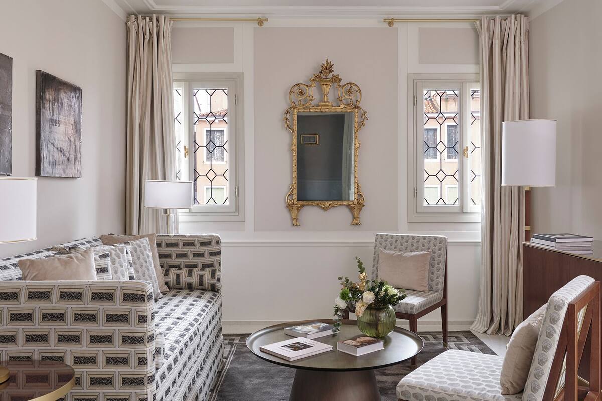 A room with a patterned sofa, a round coffee table with books and a vase, a chair, and a large ornate mirror.