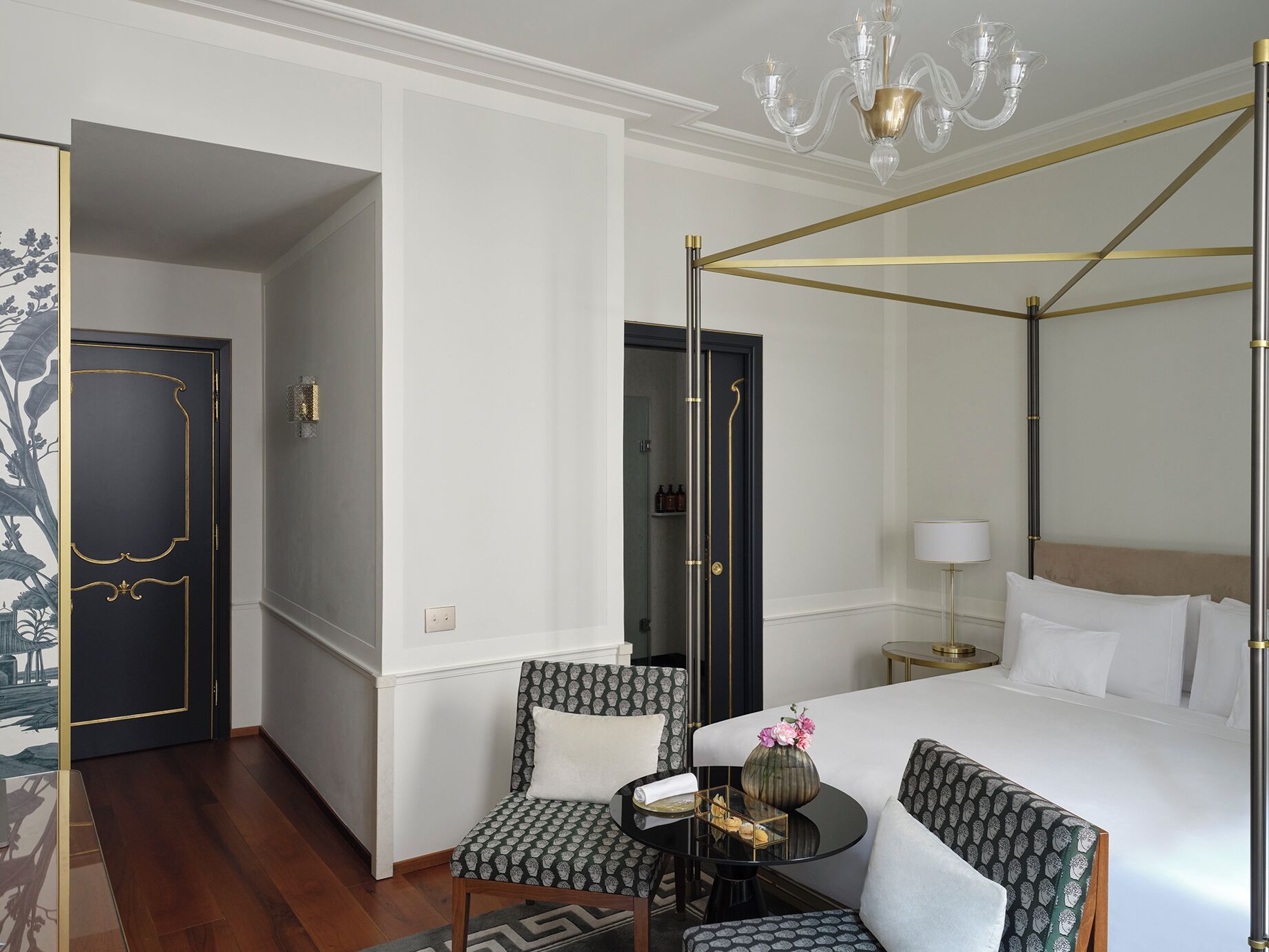 A hotel room with a four-poster bed, two patterned armchairs, a small round table with a vase and a tray, and a chandelier.