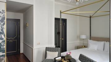 Una habitación de hotel con una cama con dosel, dos sillones estampados, una mesita redonda con un florero y una bandeja, y un candelabro.