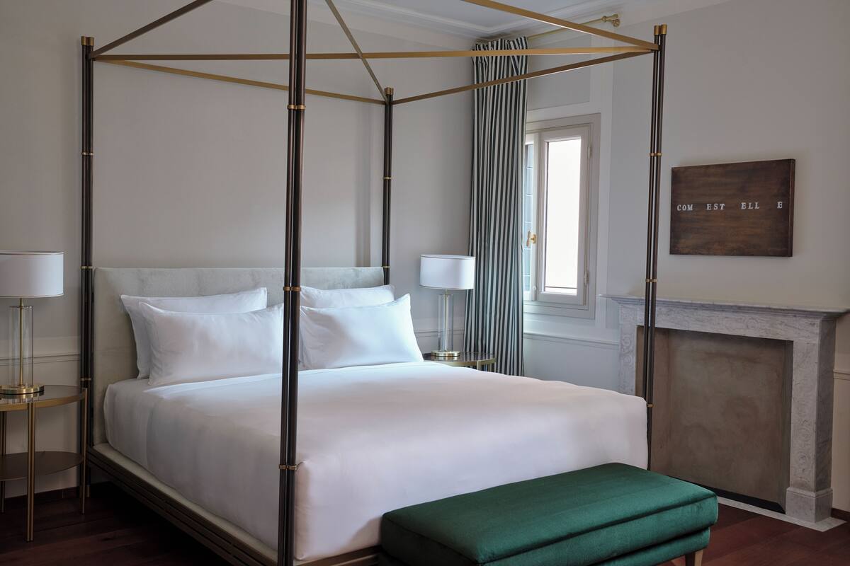 A four-poster bed with white linens, a green bench, a nightstand with a lamp, and a fireplace.