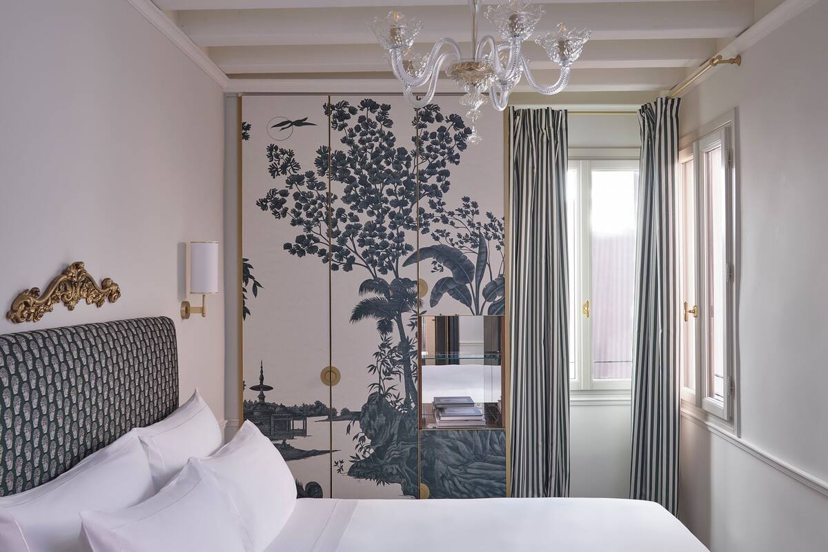 A hotel room with a bed, a chandelier, a view of a cityscape through a window, and a door with striped curtains.