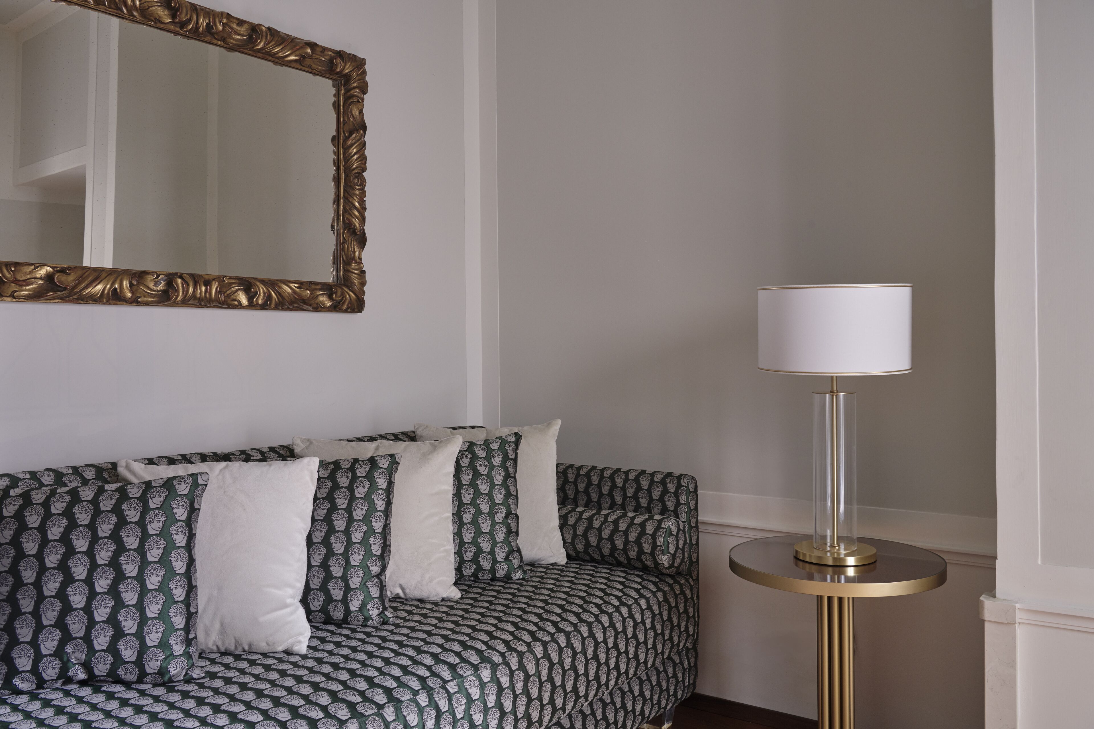 A room with a patterned sofa, a round table with a lamp, and a large mirror on the wall.