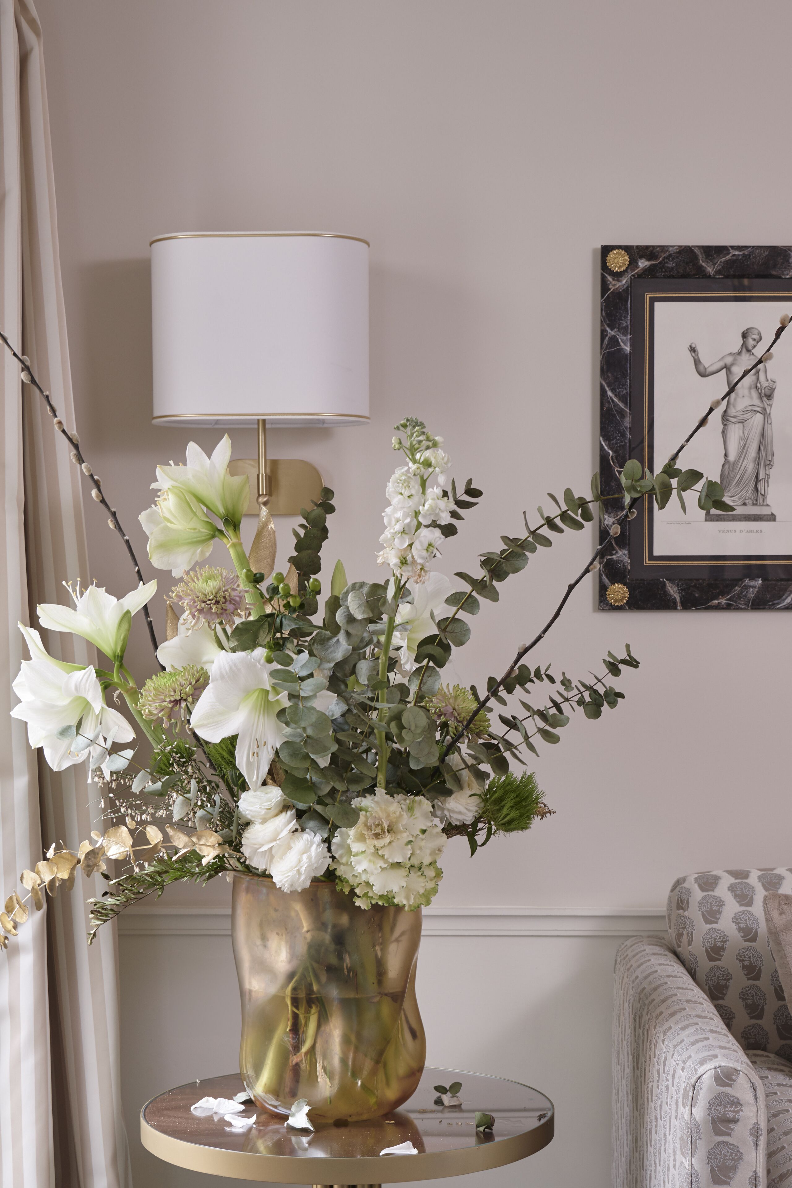Ein Zimmer mit einem runden Tisch, auf dem sich eine Vase mit weißen Blumen, eine Lampe und ein gerahmtes Bild befinden. 