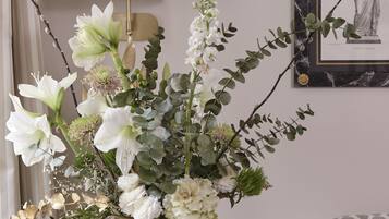 Ein Zimmer mit einem runden Tisch, auf dem sich eine Vase mit weißen Blumen, eine Lampe und ein gerahmtes Bild befinden.