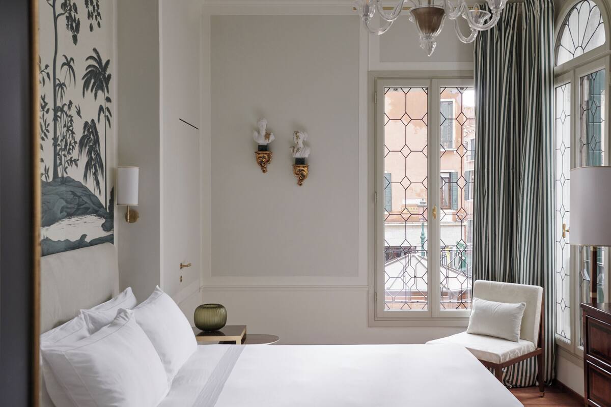 A hotel room with a bed, a chair, a nightstand, a wall-mounted lamp, and a window with curtains.