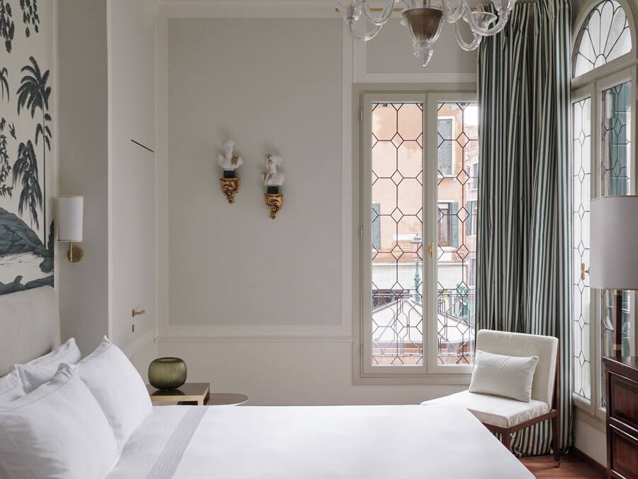 Una habitación de hotel con una cama, una silla, una mesita de noche, una lámpara de pared y una ventana con cortinas.
