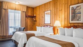 Ein Hotelzimmer mit Holzwandverkleidung, zwei Betten, einem Schreibtisch, einem Stuhl, einem fenster mit Vorhängen, einer Lampe und einem gerahmten Bild an der Wand.