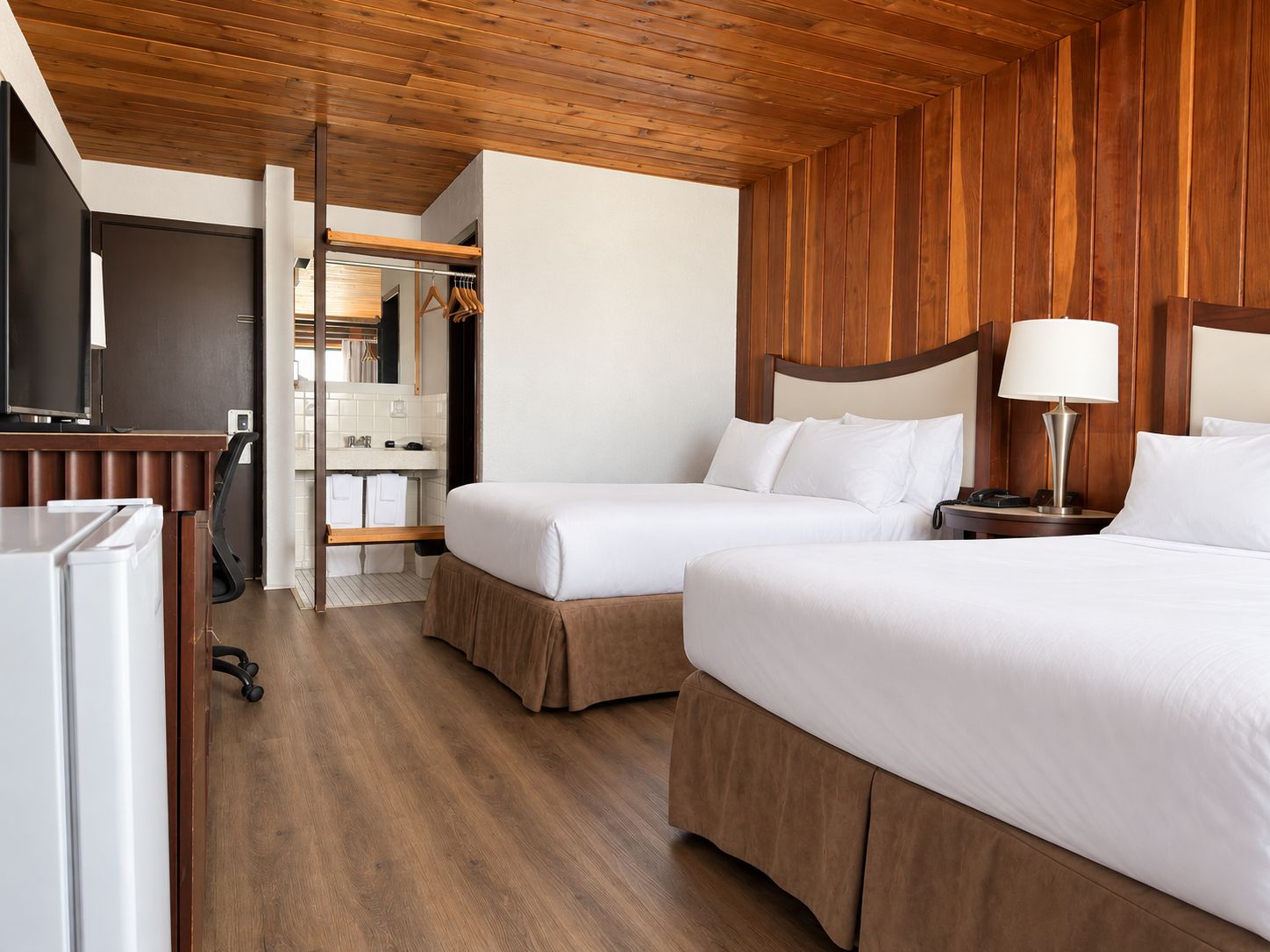Habitación de hotel con dos camas, techo de madera, un escritorio con lámpara y vista al baño.