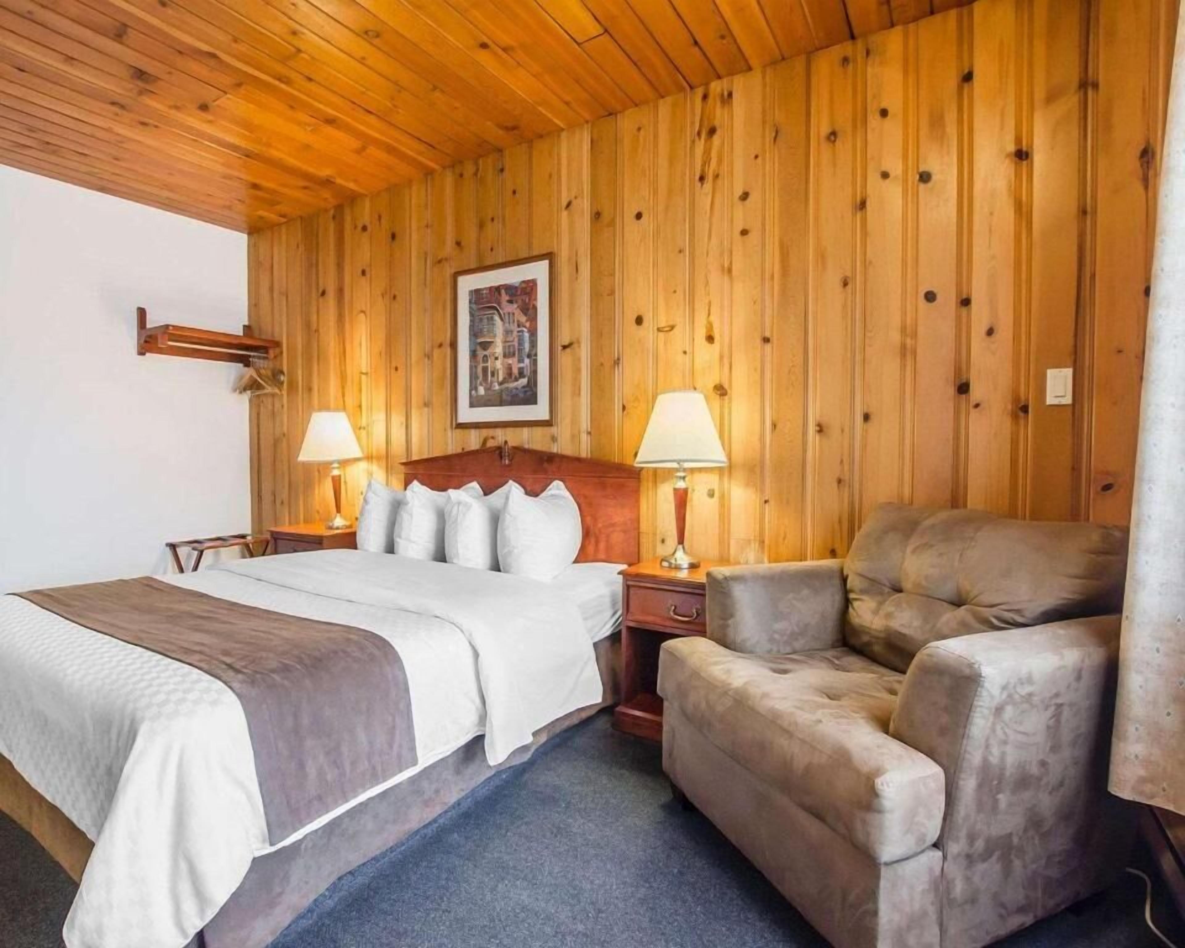 Quarto com painéis de madeira, uma cama, duas luminárias de apoio, uma cadeira e um quadro emoldurado na parede.