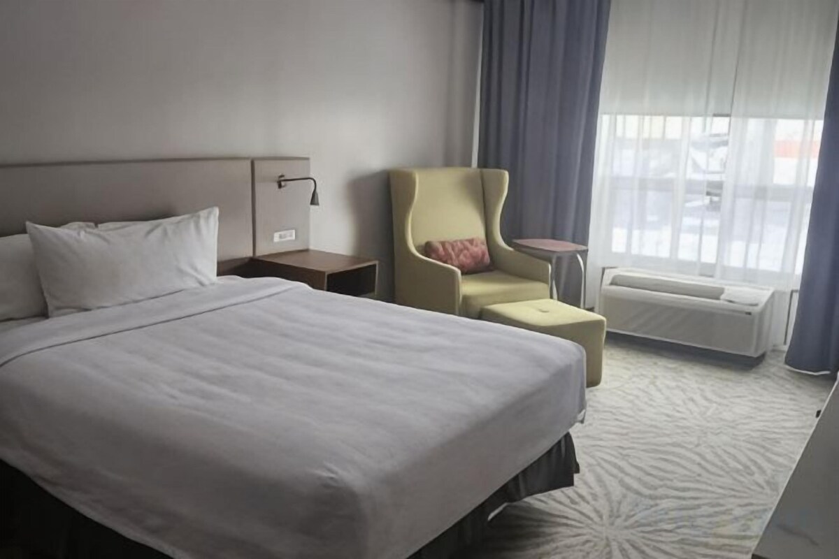 A hotel room with a bed, a chair, a small table, and a window with curtains.
