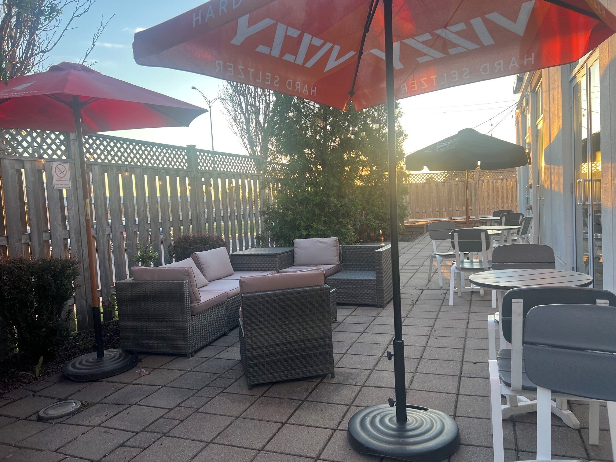En udendørs terrasse med fletmøbler, røde parasoller og et træhegn.
