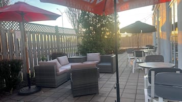 En udendørs terrasse med fletmøbler, røde parasoller og et træhegn.