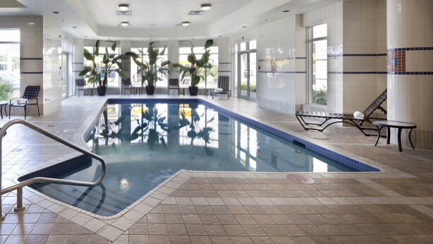 A large indoor swimming pool with clear water, surrounded by tiled walls and columns, with several potted plants and lounge chairs.