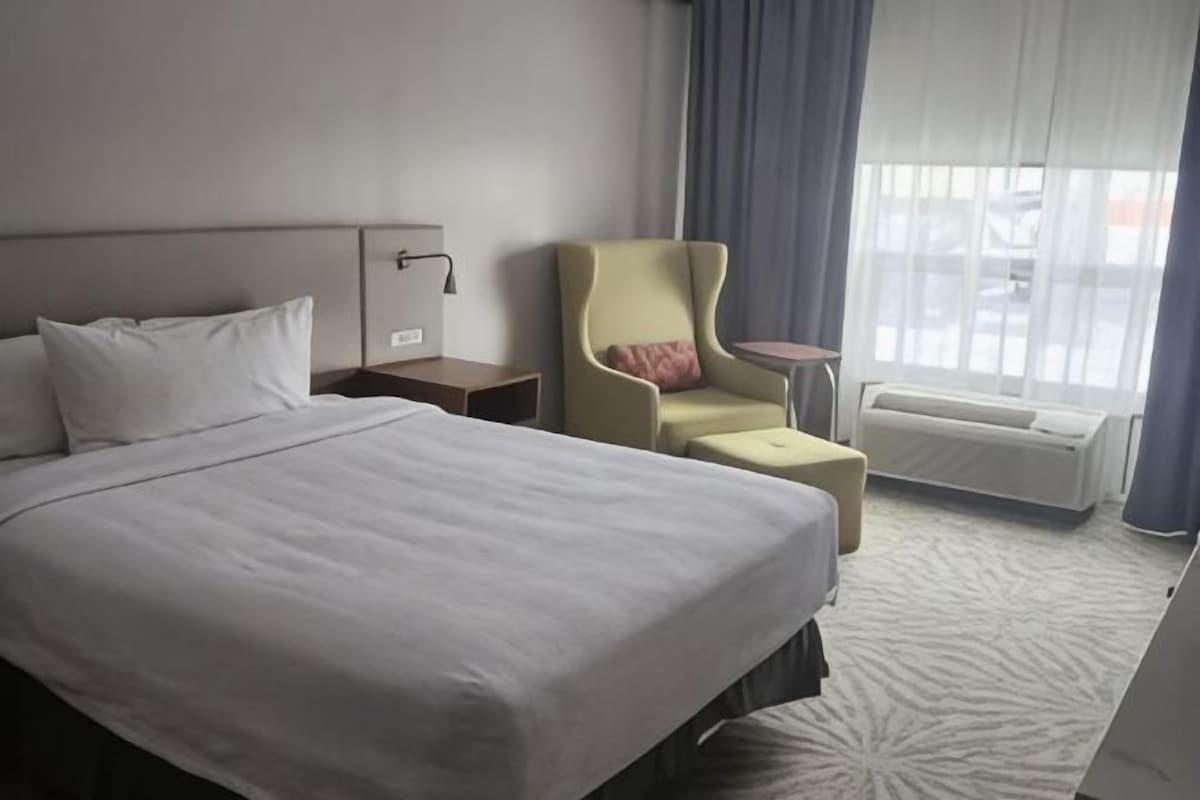 Una habitación de hotel con cama, una silla, una mesita, una mesita de noche y una ventana con cortinas.