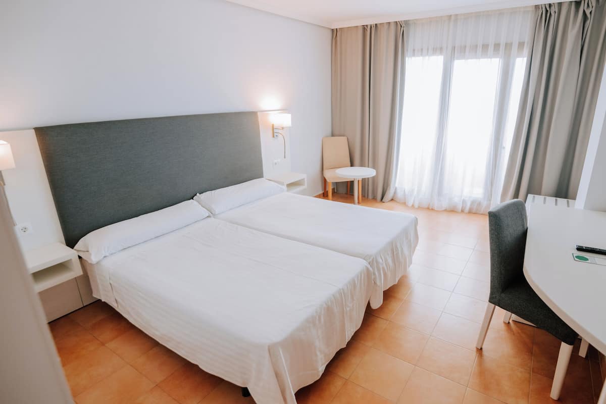 A hotel room with two beds, a desk, a chair, a TV, and a window with curtains.