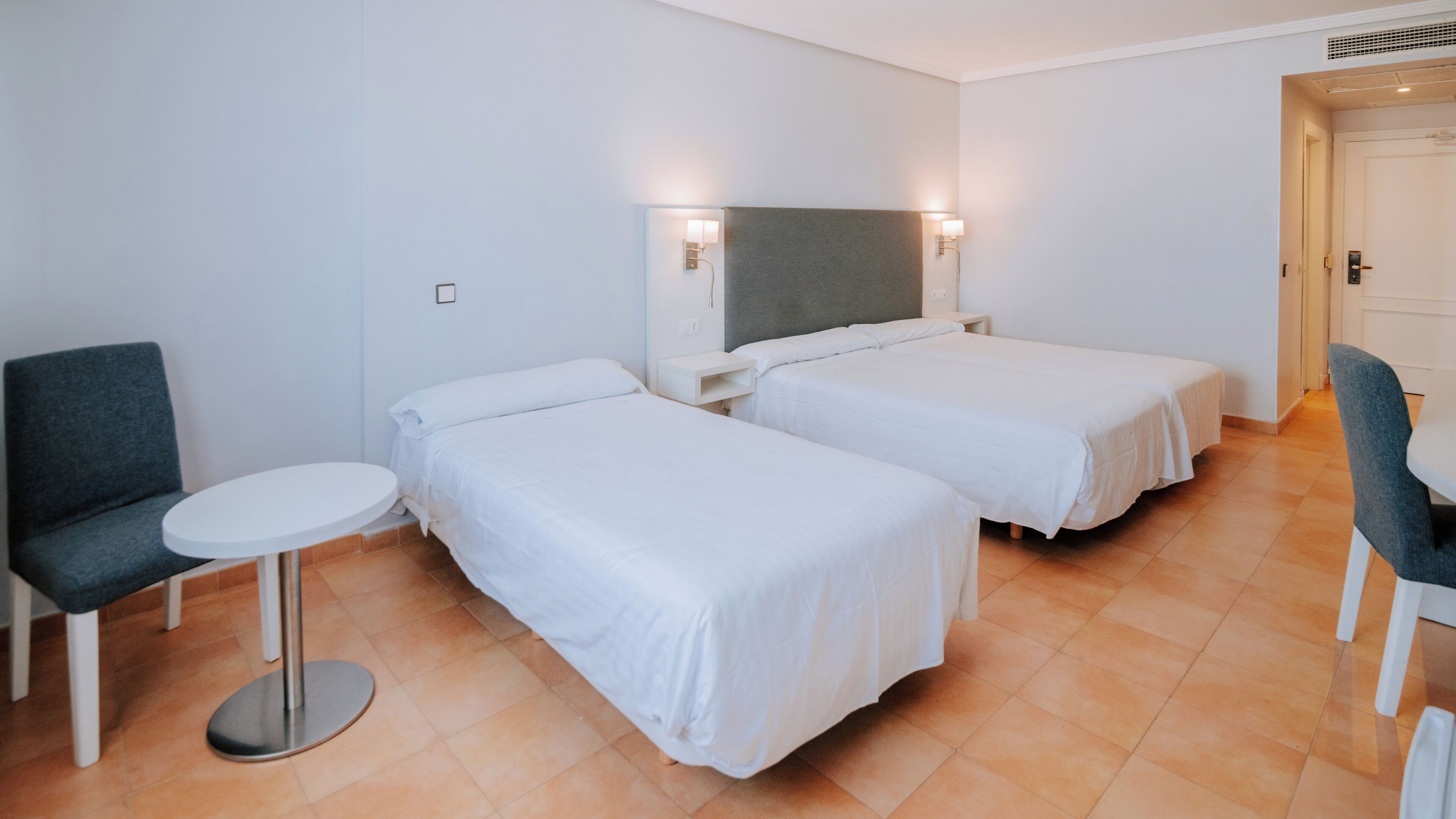 A hotel room with two single beds, a round table, a chair, and a desk.