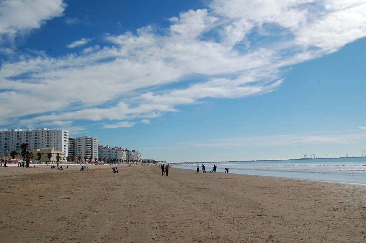 A wide sandy beach with a clear sky and a few clouds. In the background, there are several multi-story buildings, likely hotels, along the coastline.