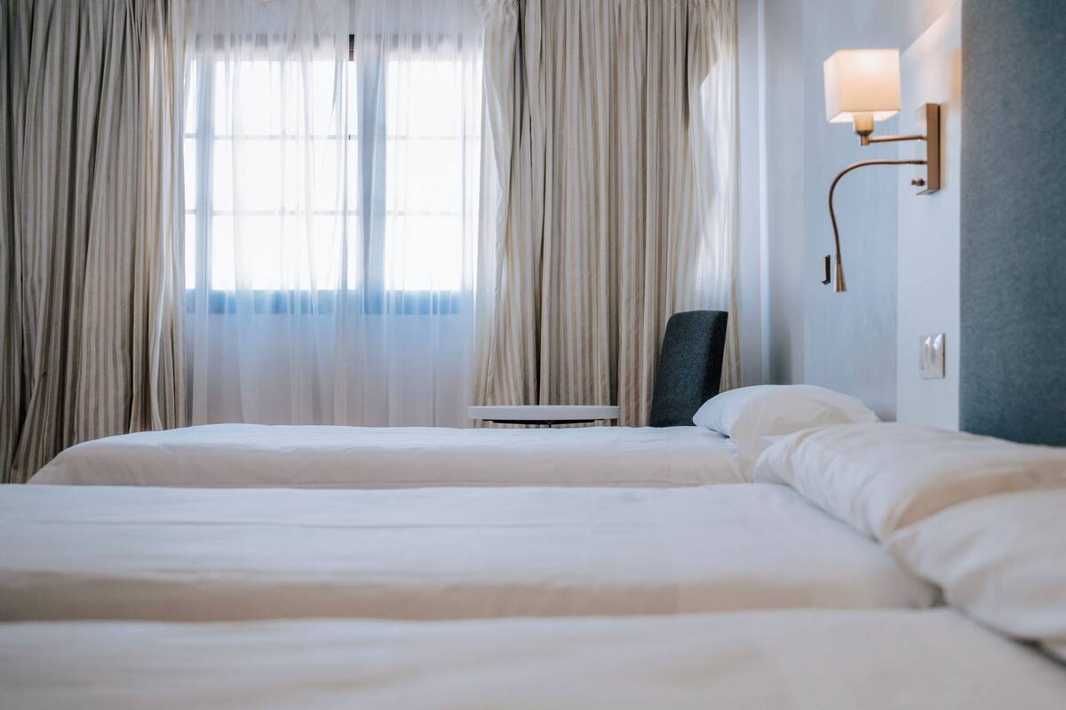 A hotel room with two beds, a chair, a lamp, and a window with curtains.