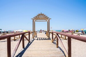 Une promenade en bois menant à une plage où l’on aperçoit des parasols et des personnes au loin.