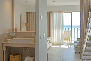 Habitación de hotel con un ventanal grande, balcón, cama, sofá y baño con lavamanos y espejo.