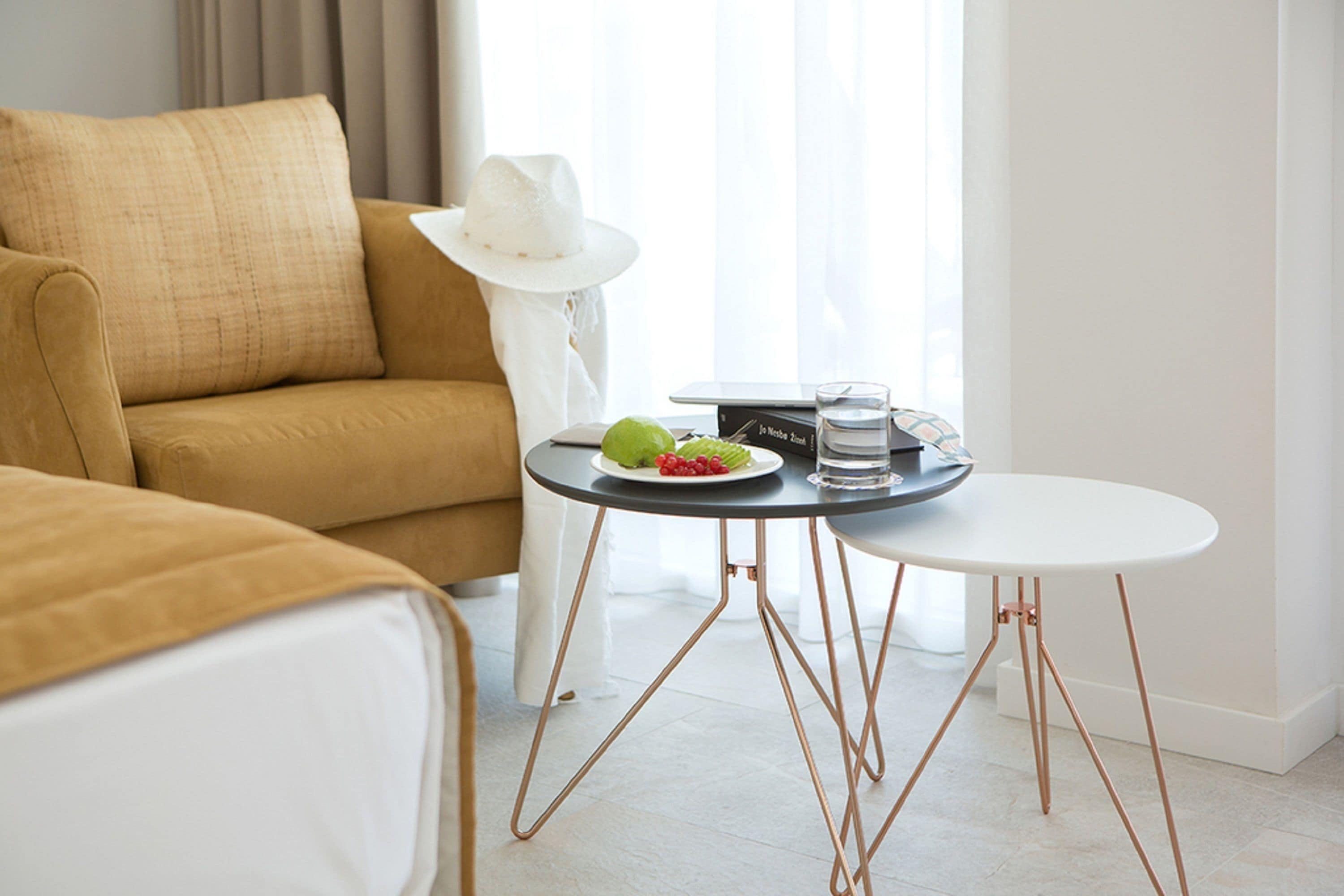 A modern hotel room with a brown sofa, a small round table with a laptop, a plate of fruit, and a white hat on the sofa.