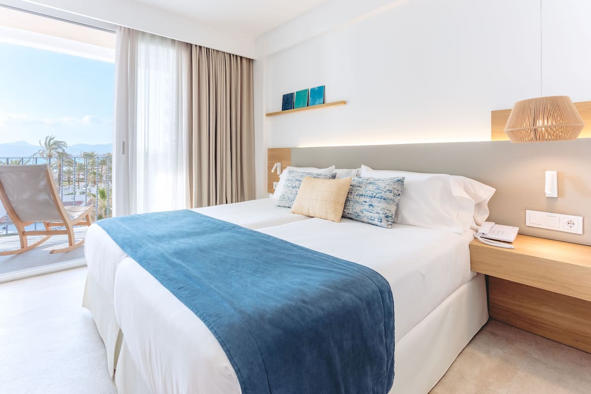A modern hotel room with a large bed, a wooden nightstand, a desk, and a view of palm trees and mountains.