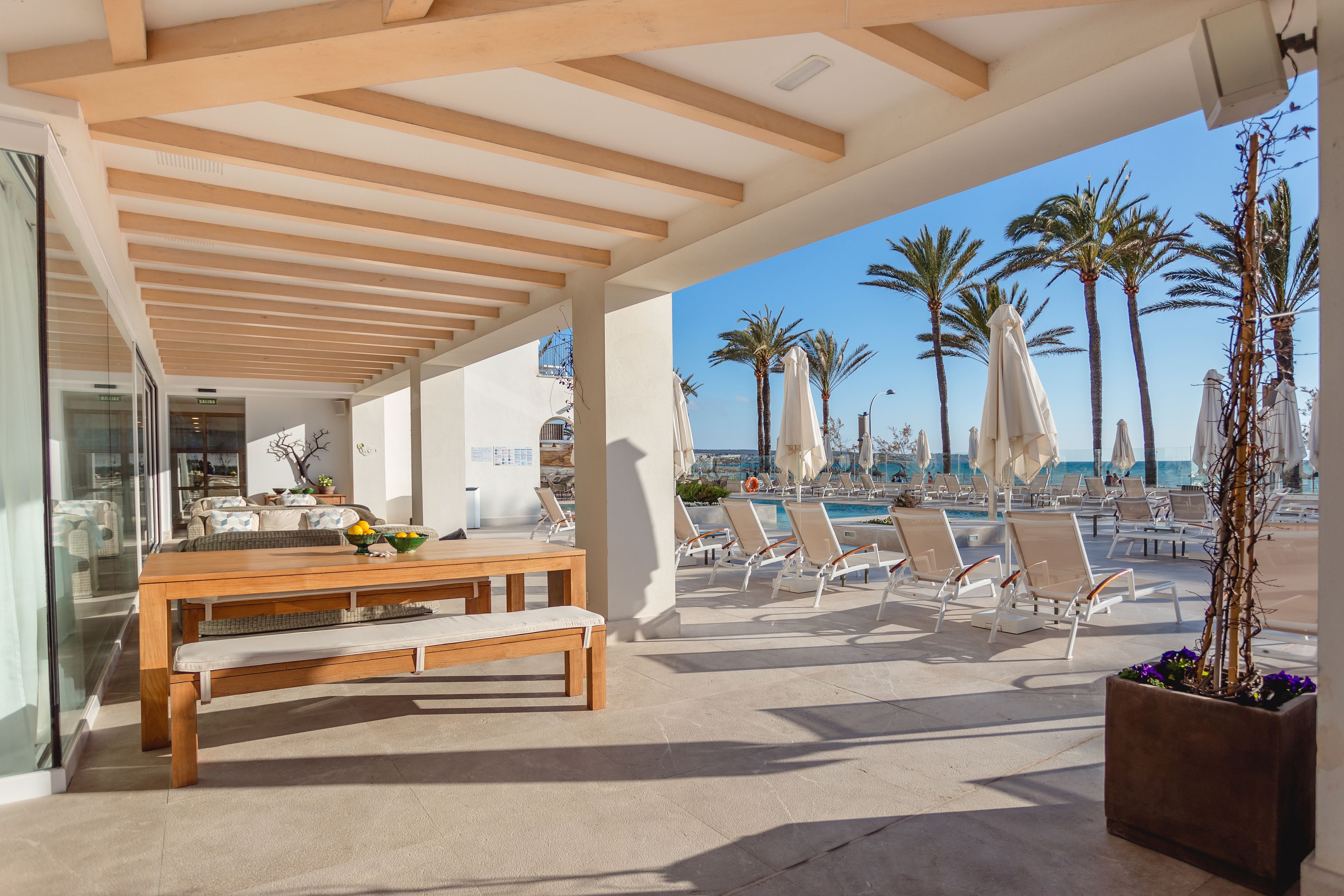 A covered outdoor seating area with a wooden table and benches, overlooking a pool with lounge chairs and palm trees.