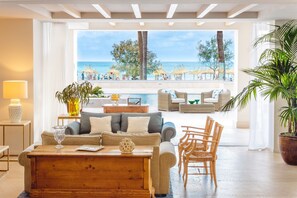 Amplia sala de estar con un gran ventanal con vista a la playa, amueblada con un sofá, sillas y una mesa de centro de madera.