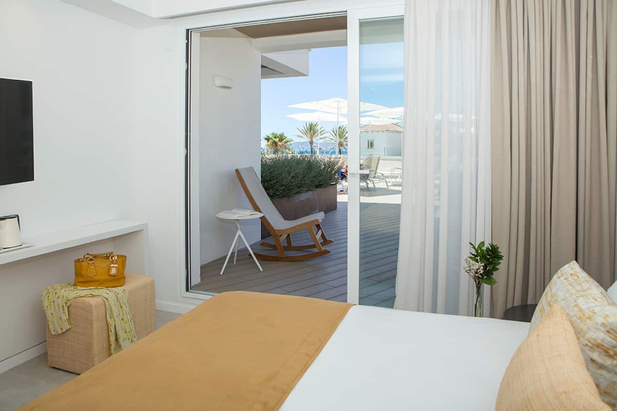 A hotel room with a bed, a small table, a rocking chair, and a view of the outdoor area with umbrellas.
