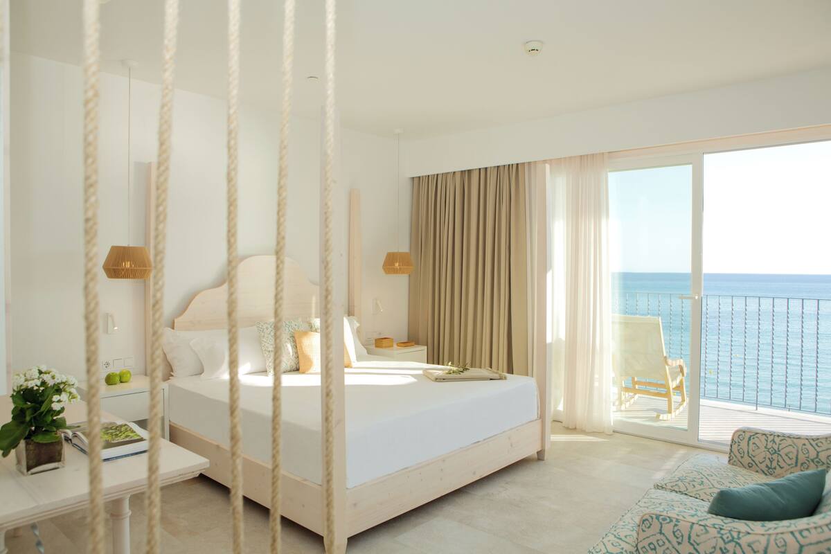 A bedroom with a large bed, a sofa, a balcony with a view of the sea, and a plant on the bedside table.