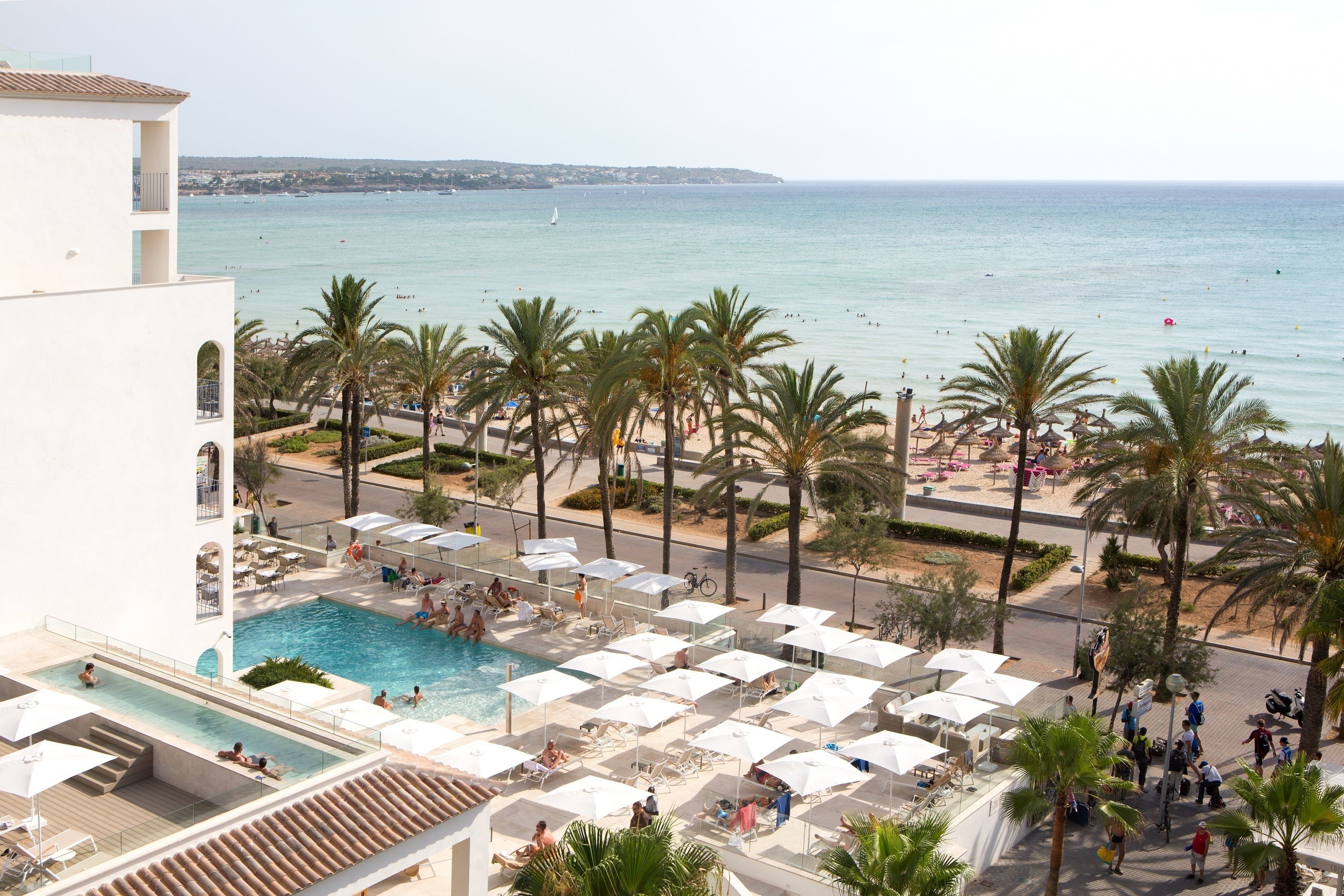 Un espace piscine aménagé dans un complexe hôtelier, avec des chaises longues et des parasols, une plage en arrière-plan et des palmiers.