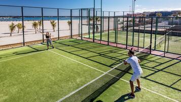 Un court de tennis avec un filet, entouré d’une clôture, et une plage en arrière-plan.