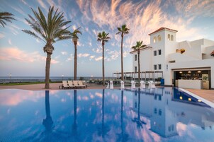 Un área de piscina con sillones de descanso, palmeras y un edificio de fachada blanca y una torre.