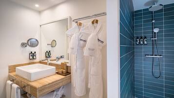 A modern bathroom with a white sink, wooden vanity, and a large mirror. There are white robes hanging on a rack and a shower with a handheld showerhead.