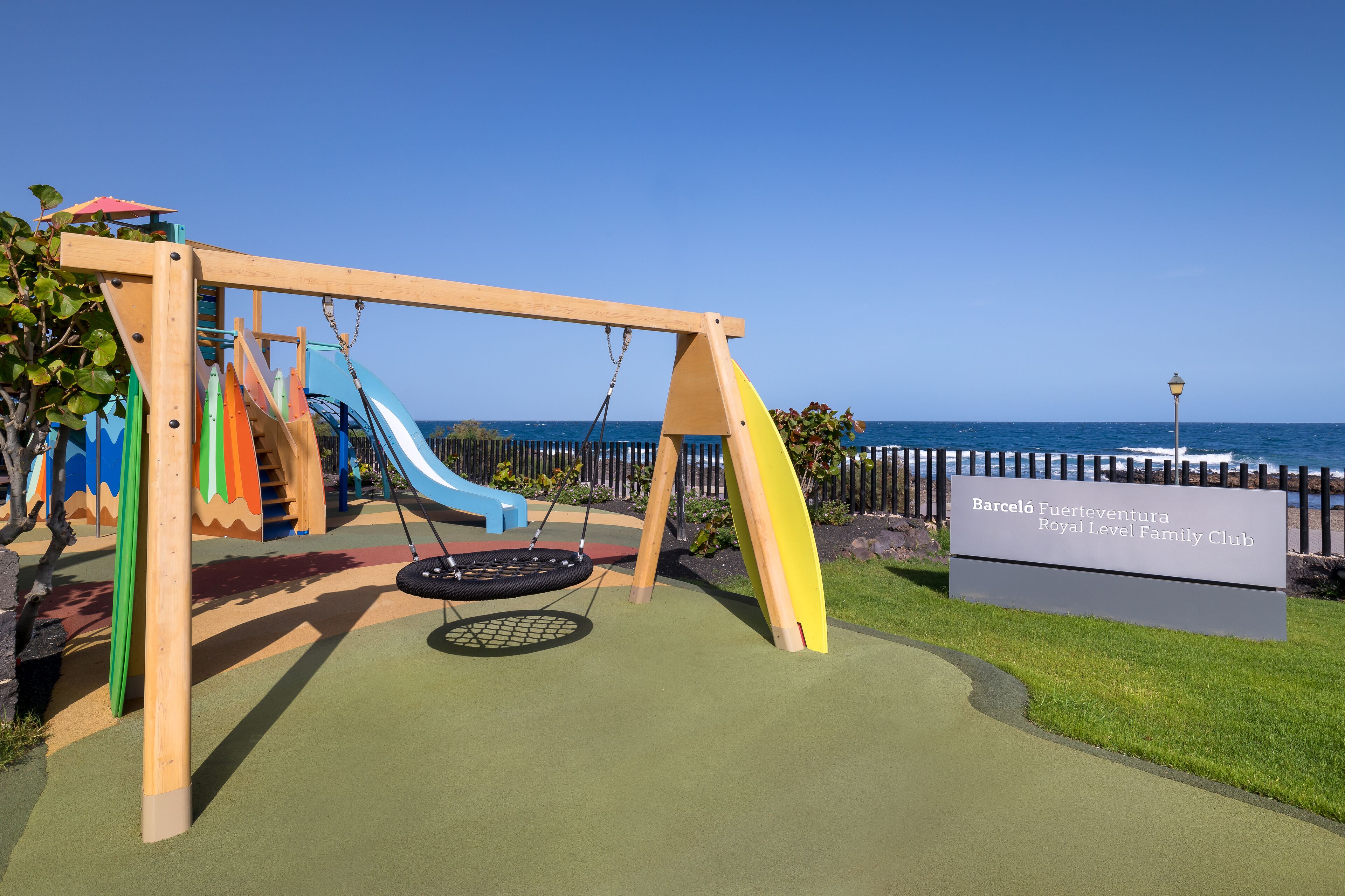 Een speeltuin met schommels en glijbanen, een bord met daarop Barceló Fuertesventura Royal Level Family Club, en uitzicht op de zee.
