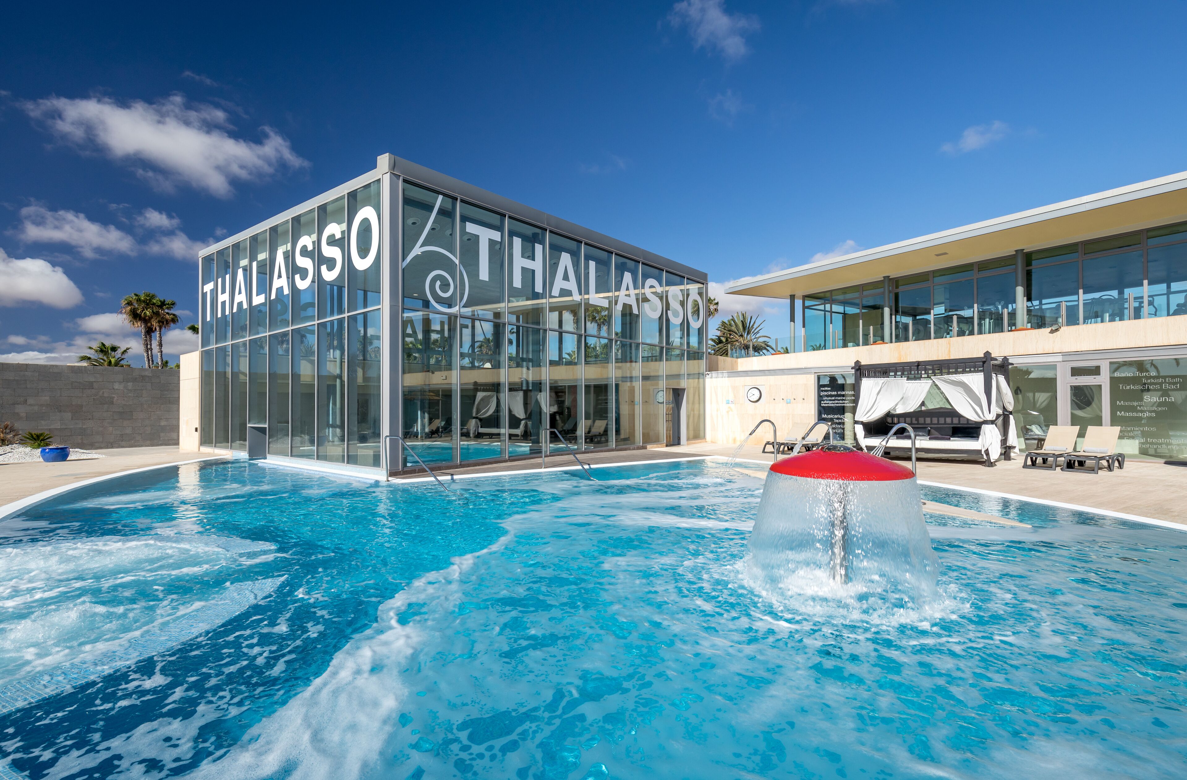A modern building with a glass facade and the name "THALASSO" prominently displayed. In front of the building is a swimming pool with clear blue water and a water feature.
