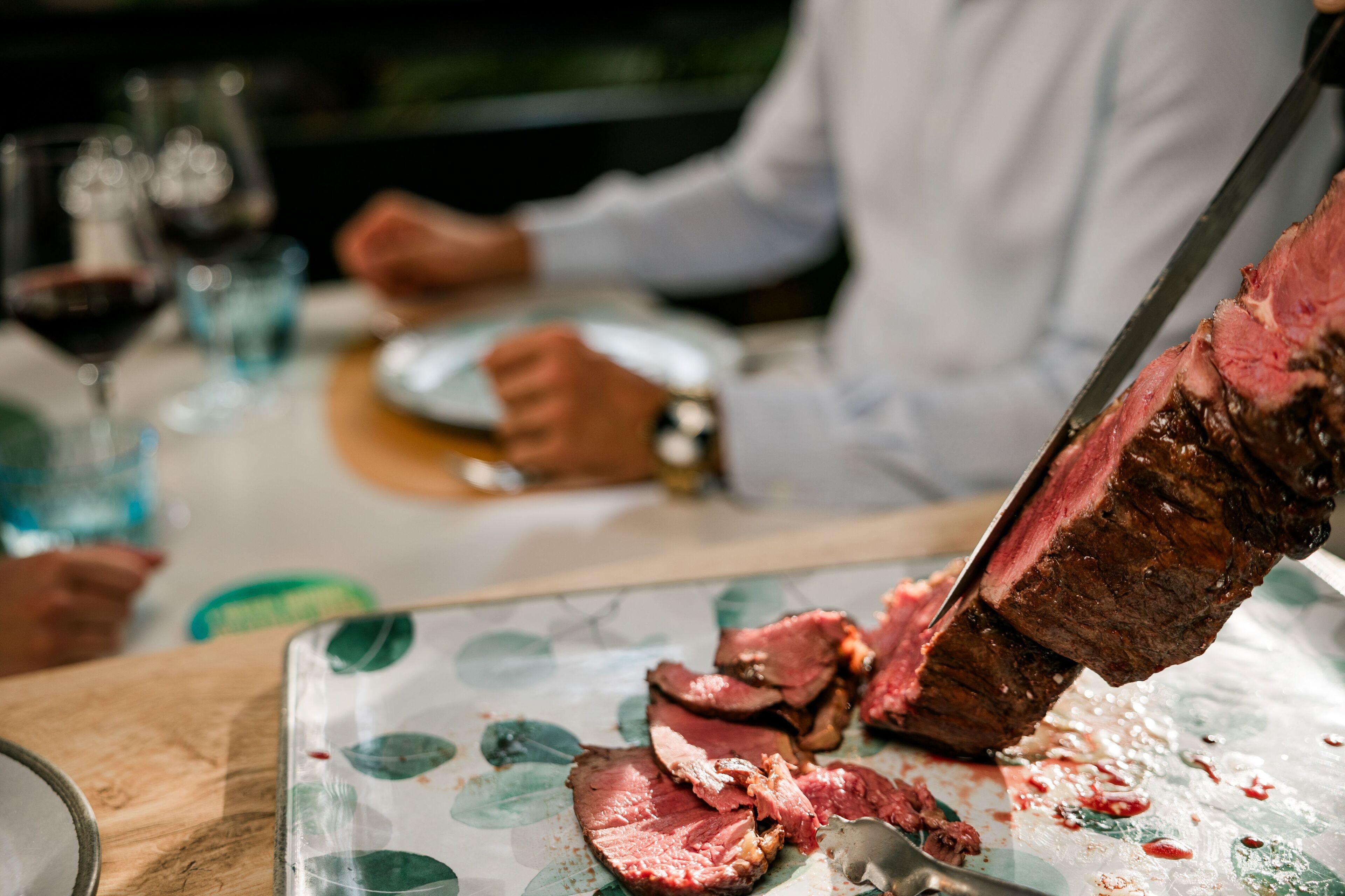 Una mesa con un gran trozo de carne siendo cortado, copas de vino tinto y una persona al fondo.