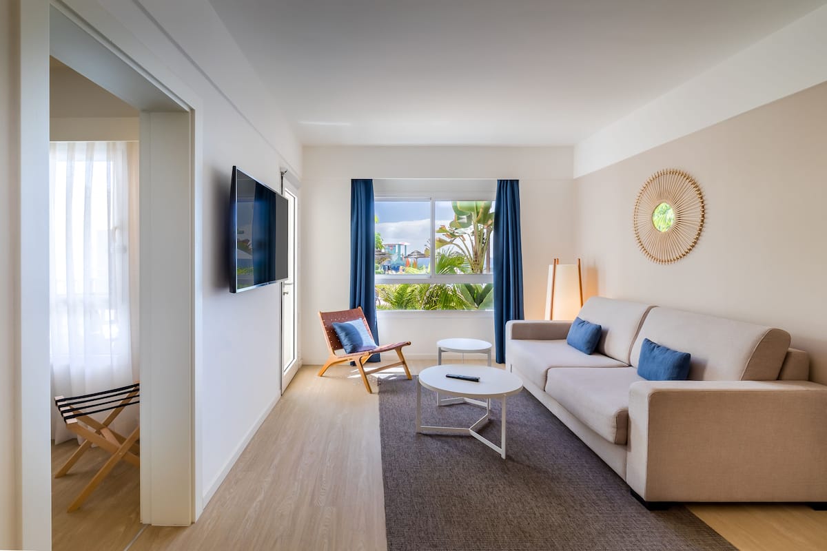 Ein modernes Wohnzimmer mit Sofa, Sessel und Couchtisch, einem Flachbildfernseher und einem großen Fenster mit Vorhängen.