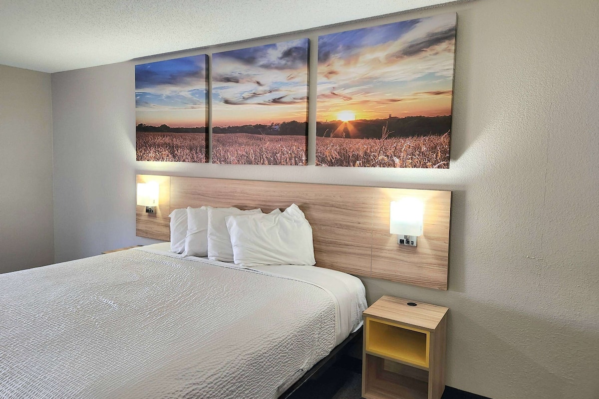 Hotellihuone, jossa on suuri sänky, puinen pääty, kaksi yöpöytävalaisinta ja seinälle ripustettu maalaus, jossa on kuvattu auringonlasku pellon yllä.