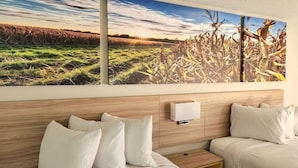 Ein Hotelzimmer mit großem Landschaftsbild, einem Holzkopfteil, zwei Betten mit weißer Bettwäsche und einem Nachttisch mit Lampe.