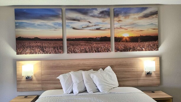 Ein Hotelzimmer mit großem Wandbild eines Sonnenuntergangs, einem Holzkopfteil, weißer Bettwäsche und Nachttischlampen.