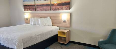 Una habitación de hotel con una cama, una mesita de noche con lámpara, una silla y un cuadro en la pared que representa un atardecer sobre un campo.