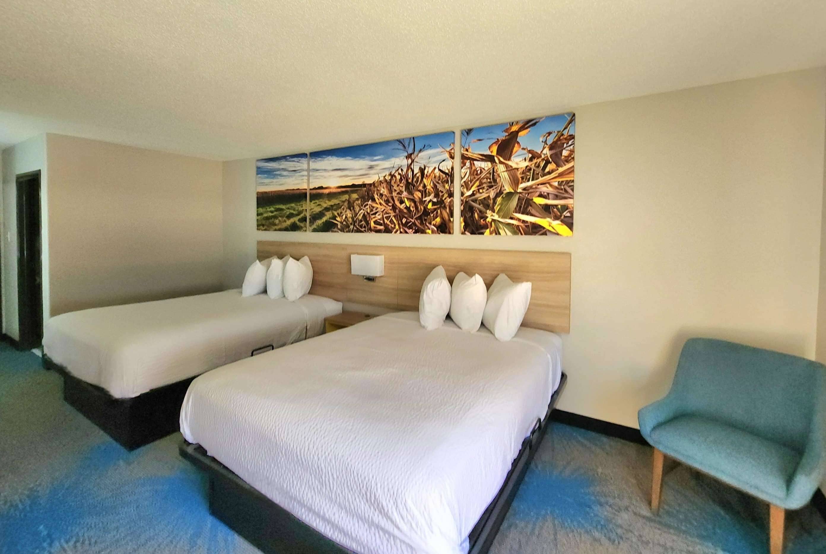 Una camera d'albergo con due letti, un grande dipinto che raffigura un paesaggio e una poltrona blu.