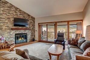 Ein Wohnzimmer mit Kamin aus Stein, einem Flachbildfernseher, einer Couch, einem Couchtisch und Blick auf einen Balkon mit einer schönen Landschaft.