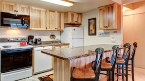 A kitchen with wooden cabinets, a white refrigerator, a stove, and a coffee maker. There are four bar stools with cushioned seats. - Trails End Condominiums by Great Western Lodging (Breckenridge)