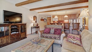 A living room with a television, wooden cabinet, patterned sofa, and a dining area with chairs and a table. - Trails End Condominiums by Great Western Lodging (Breckenridge)