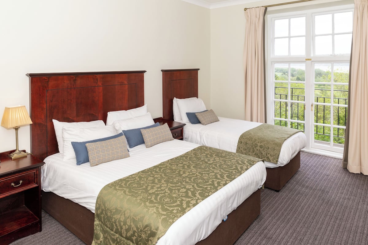 A hotel room with two beds, a nightstand, a lamp, and a large window with a view of greenery.