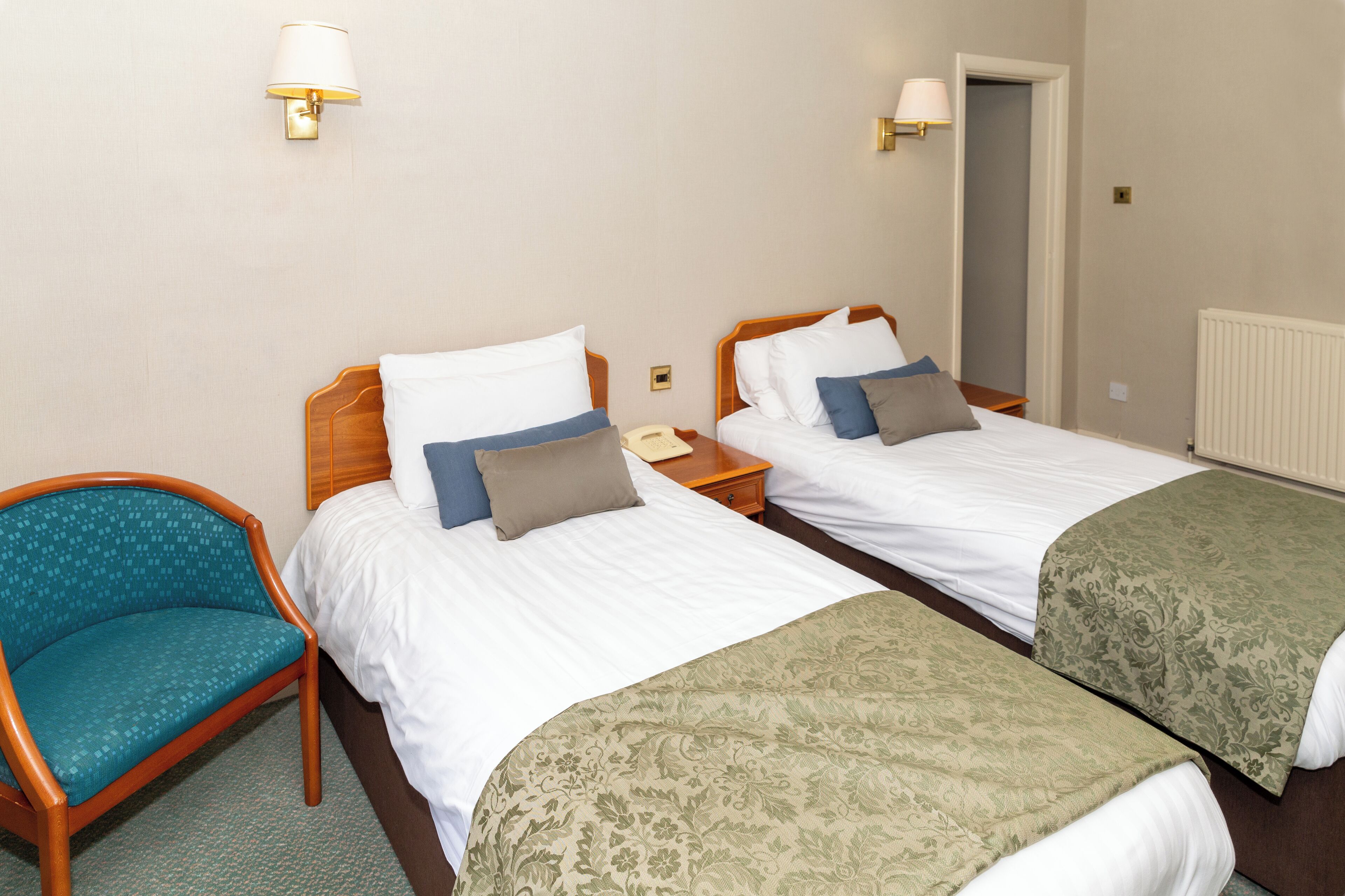 A hotel room with two single beds, a wooden nightstand, a teal armchair, and a door leading to another room.