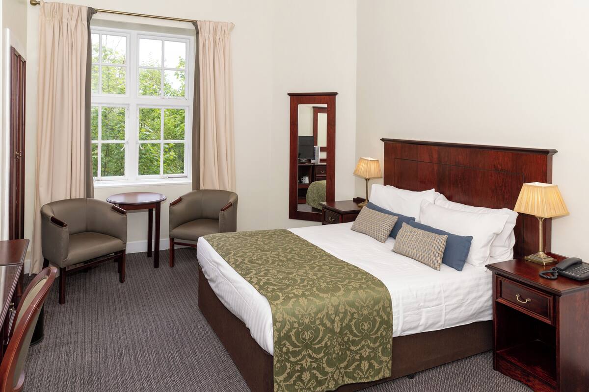 A hotel room with a large bed, two armchairs, a small table, a nightstand with a lamp, and a mirror.