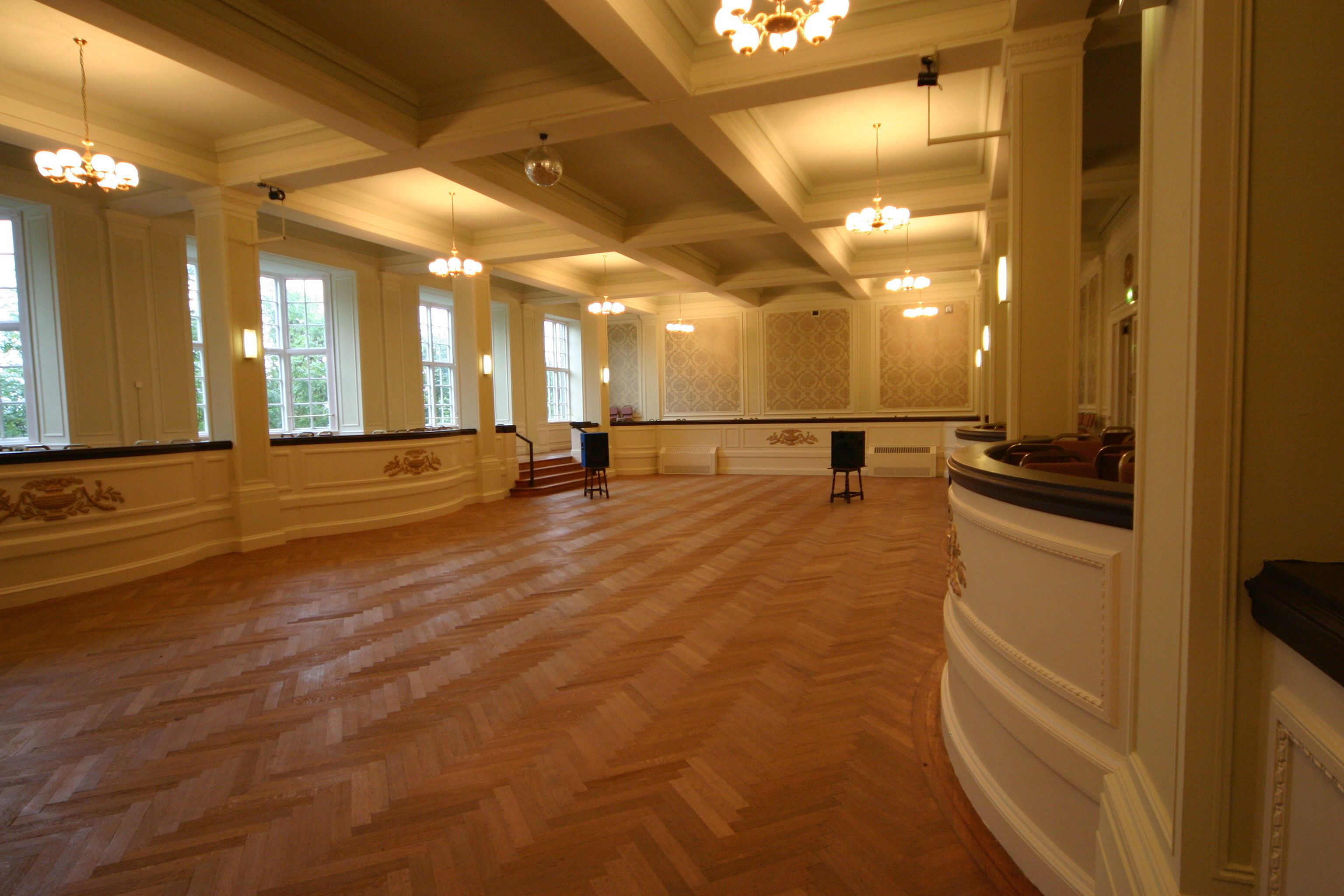 A spacious hall with wooden parquet flooring, large windows, and ornate chandeliers.